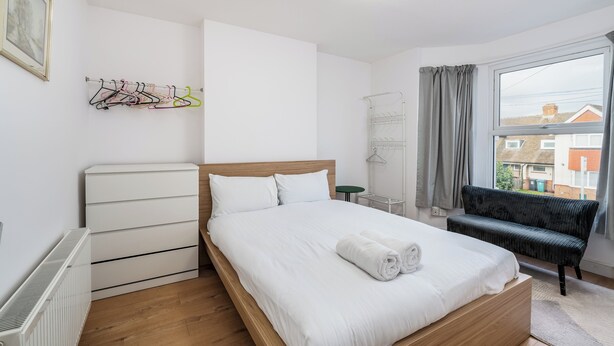 A neatly arranged bedroom with a bed, a radiator, a bench, and a window with curtains.