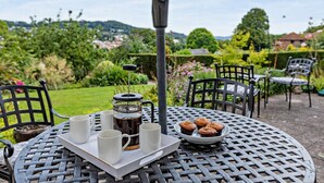 Een ronde metalen tafel met een dienblad waarop witte kopjes, een French press, een bord muffins en een metalen paraplu staan.