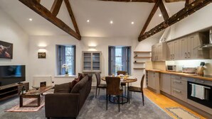 Un salon moderne avec un canapé marron, une table à manger avec des chaises, un coin cuisine avec des placards et des appareils électroménagers, et un téléviseur placé à gauche.