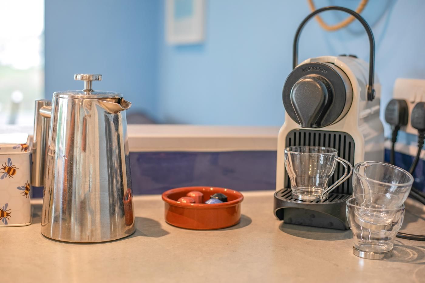 A kitchen counter with a coffee maker, a kettle, a bowl with tea bags, and two glasses.