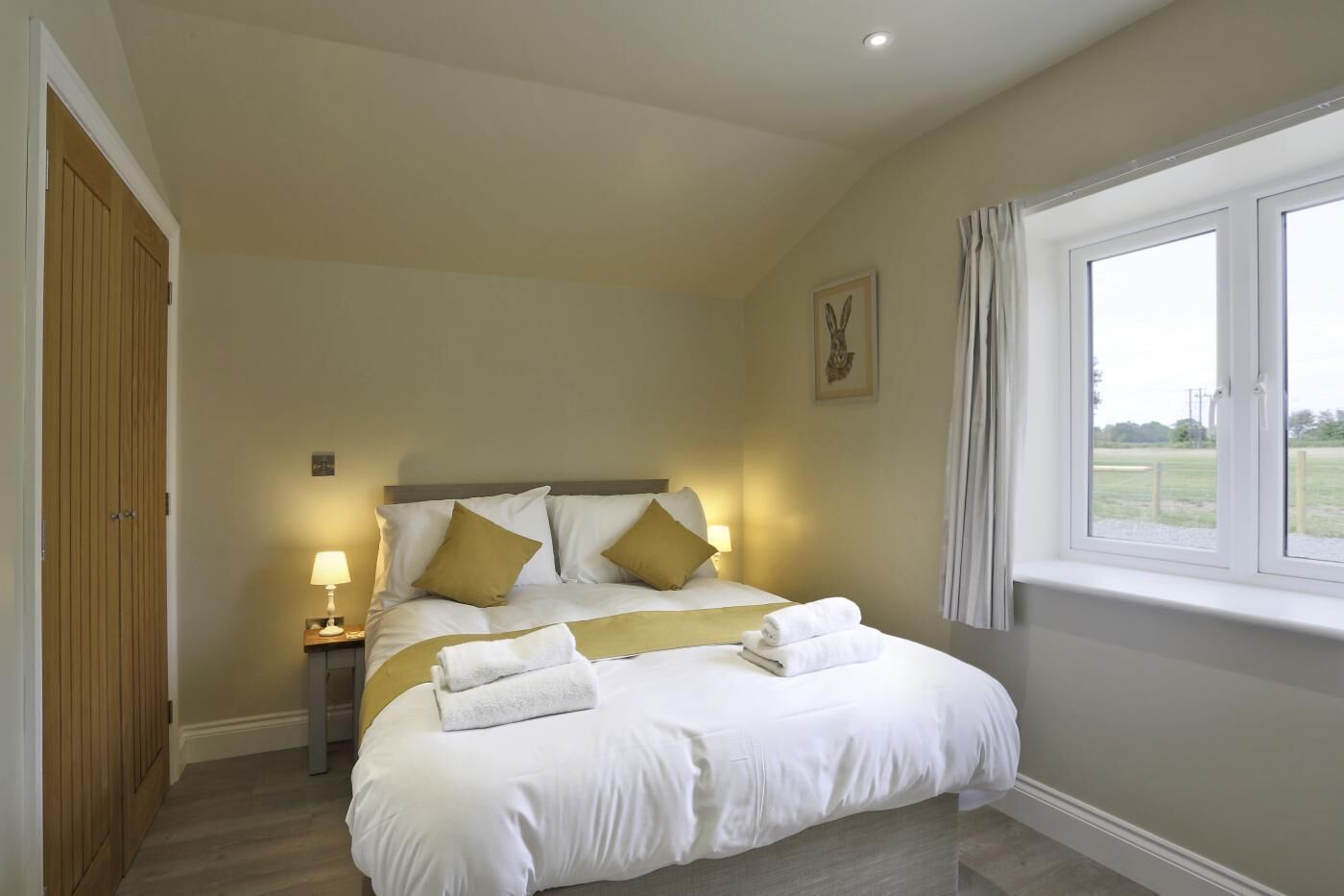 A neatly arranged bedroom with a bed, bedside lamps, a window with curtains, and a picture on the wall.