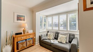 A living room with a grey sofa, a wooden side table with a lamp, a framed picture on the wall, and a window with blinds. - 4 Bed in Seahouses (oc-o27867) (Seahouses)