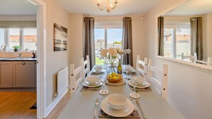 A dining room with a long table set for six, white chairs, a chandelier, and a view of the outside. - 4 Bed in Seahouses (oc-o27867) (Seahouses)