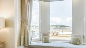 A room with a bay window overlooking a beach, featuring a white cushioned bench with two pillows, a lamp, and a view of the sea and buildings. - 9 Bed in Instow (oc-a27886) (Instow)