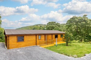 A wooden cabin with a gravel driveway, a wooden deck, and a grassy area with trees. - 3 Bed in Llanddulas (oc-r27872) (Rhyd-y-foel)