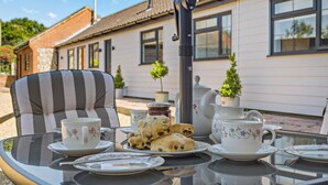 A table set for tea with floral teacups, a teapot, and scones, outside a house with a tiled roof and windows. - 2 Bed in Gimingham (oc-h27869) (Gimingham)