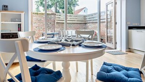 A dining area with a round table set for four, white chairs, and a view of a brick wall and trees outside. - 2 Bed in Southwold (oc-t27861) (Southwold)