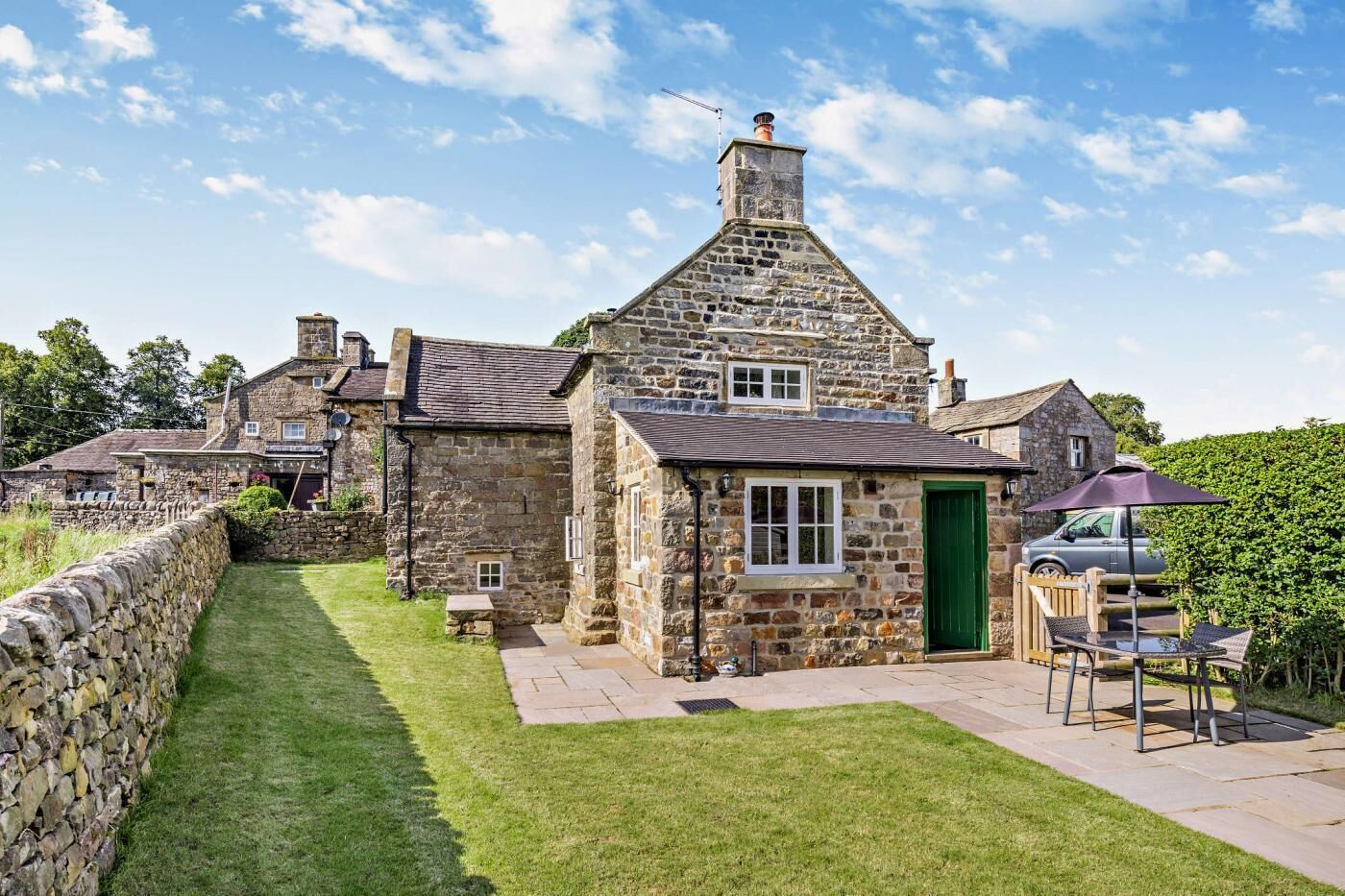 A stone cottage with a green door, a paved patio, and a car parked nearby.