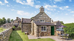 Une petite maison en pierre avec une porte verte, une terrasse dallée et une voiture garée à proximité.