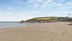 A wide sandy beach with gentle waves, a rocky outcrop, and a hill with buildings in the background. - 3 Bed in Broad Haven (oc-p27926) (Broad Haven)