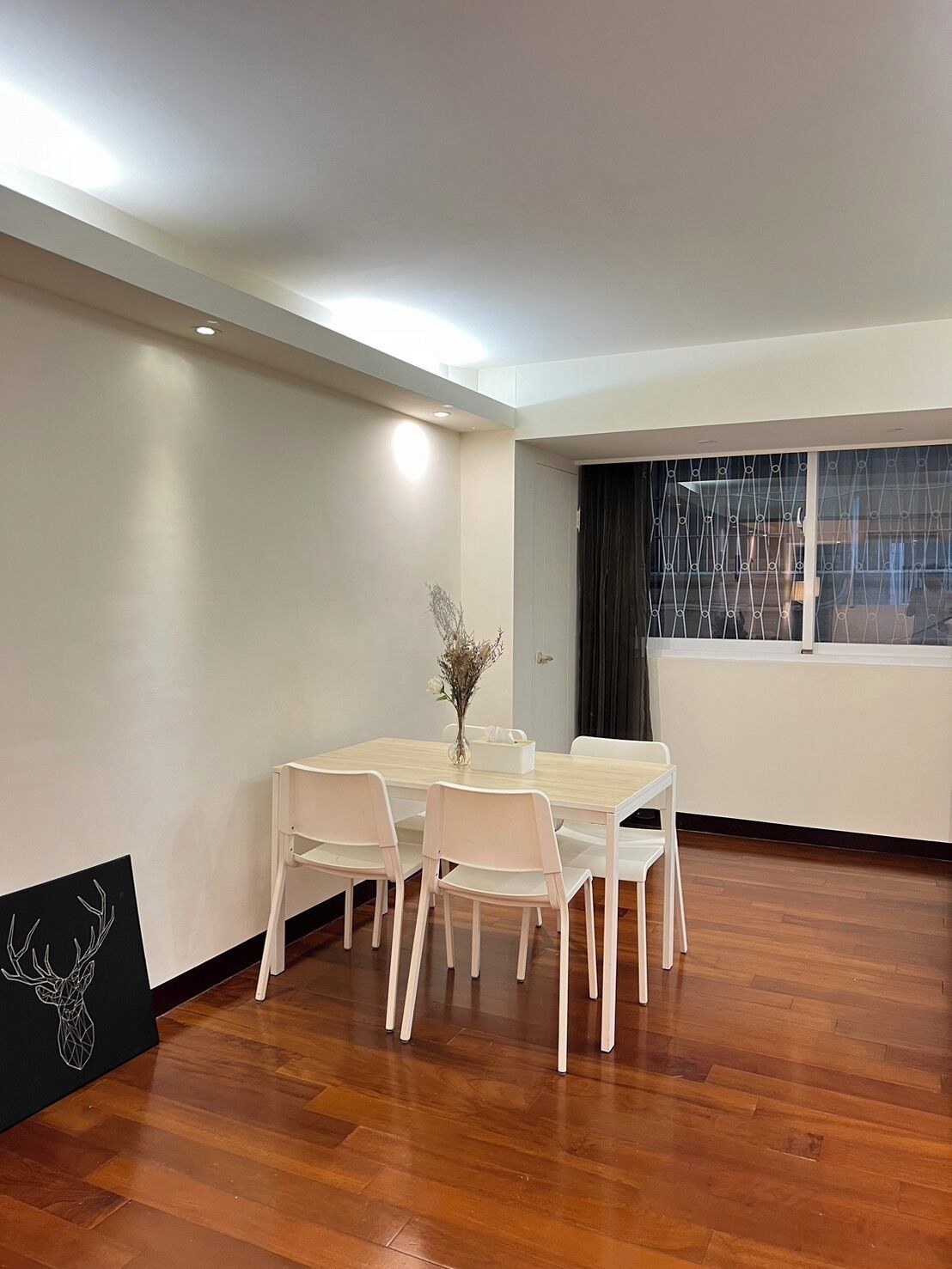 A dining area with a wooden table and white chairs, a vase with flowers, and a window with curtains.
