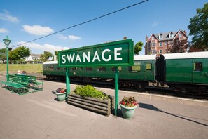 Et grønt skilt med teksten "SWANAGE", en bænk, et tog og en bygning i baggrunden.