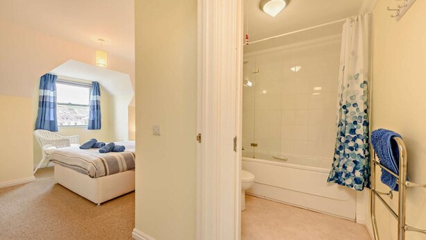 A bedroom with a bed, a window with curtains, and a bathroom with a shower, toilet, and towel rack. - 2 Bed in Broad Haven (oc-s27956) (Broad Haven)