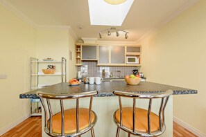 A kitchen with a central island, two bar stools, a microwave, and a bowl of fruit. - 2 Bed in Broad Haven (oc-s27956) (Broad Haven)