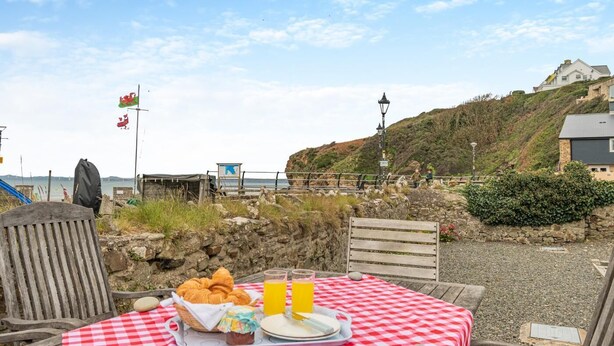 A seaside table set with croissants, a basket, and a bottle of orange juice, overlooking a rocky shoreline with a flag and a lamp post. - 4 Bed in Little Haven (oc-b27961) (Little Haven)