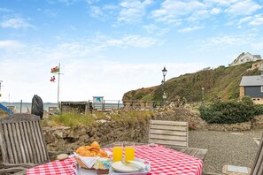 A seaside table set with croissants, a basket, and a bottle of orange juice, overlooking a rocky shoreline with a flag and a lamp post. - 4 Bed in Little Haven (oc-b27961) (Little Haven)