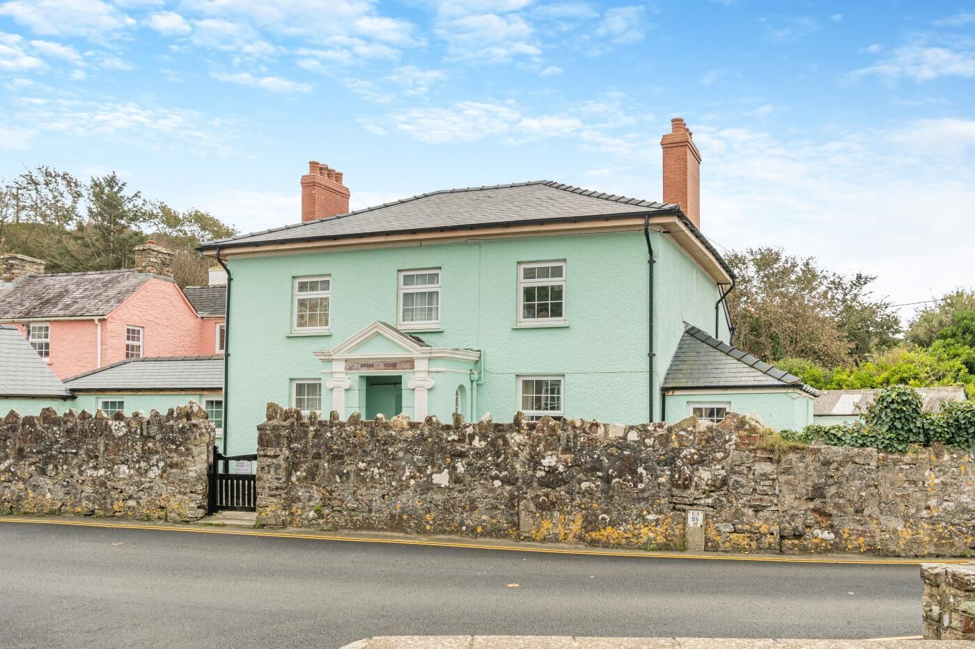Une maison à deux étages, avec un mur de pierre à l’avant, une enseigne indiquant « Bridge House », et un ciel bleu clair en arrière-plan.
