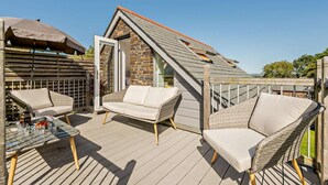 En træterrasse med fletningsmøbler, et glasbord og en bygning i sten i baggrunden.