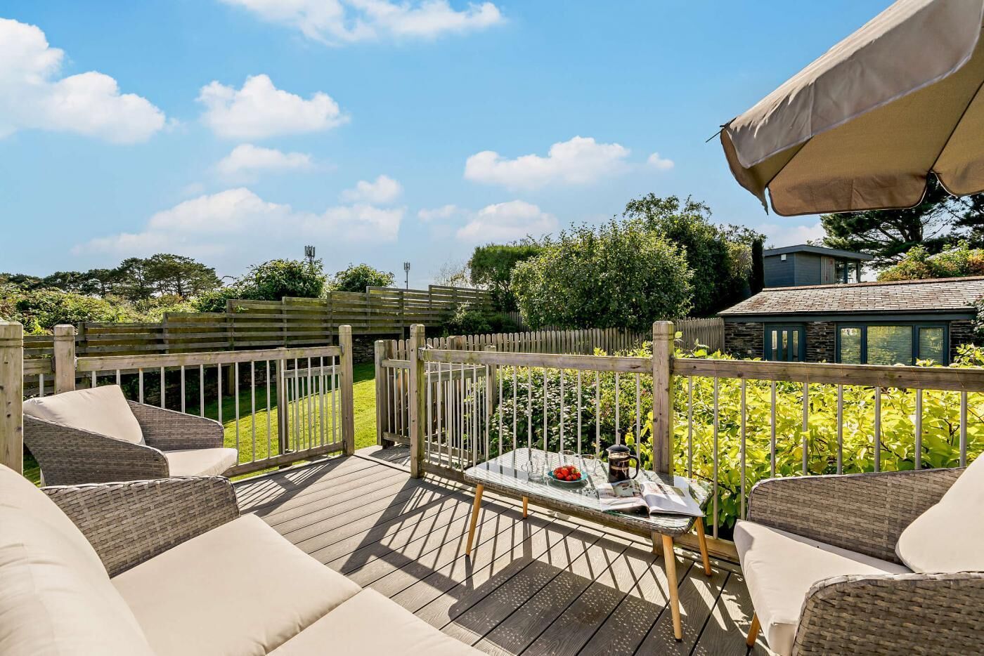 A wooden deck with outdoor furniture, a table with a pitcher and a plate of fruit, and a view of a house and greenery in the background.