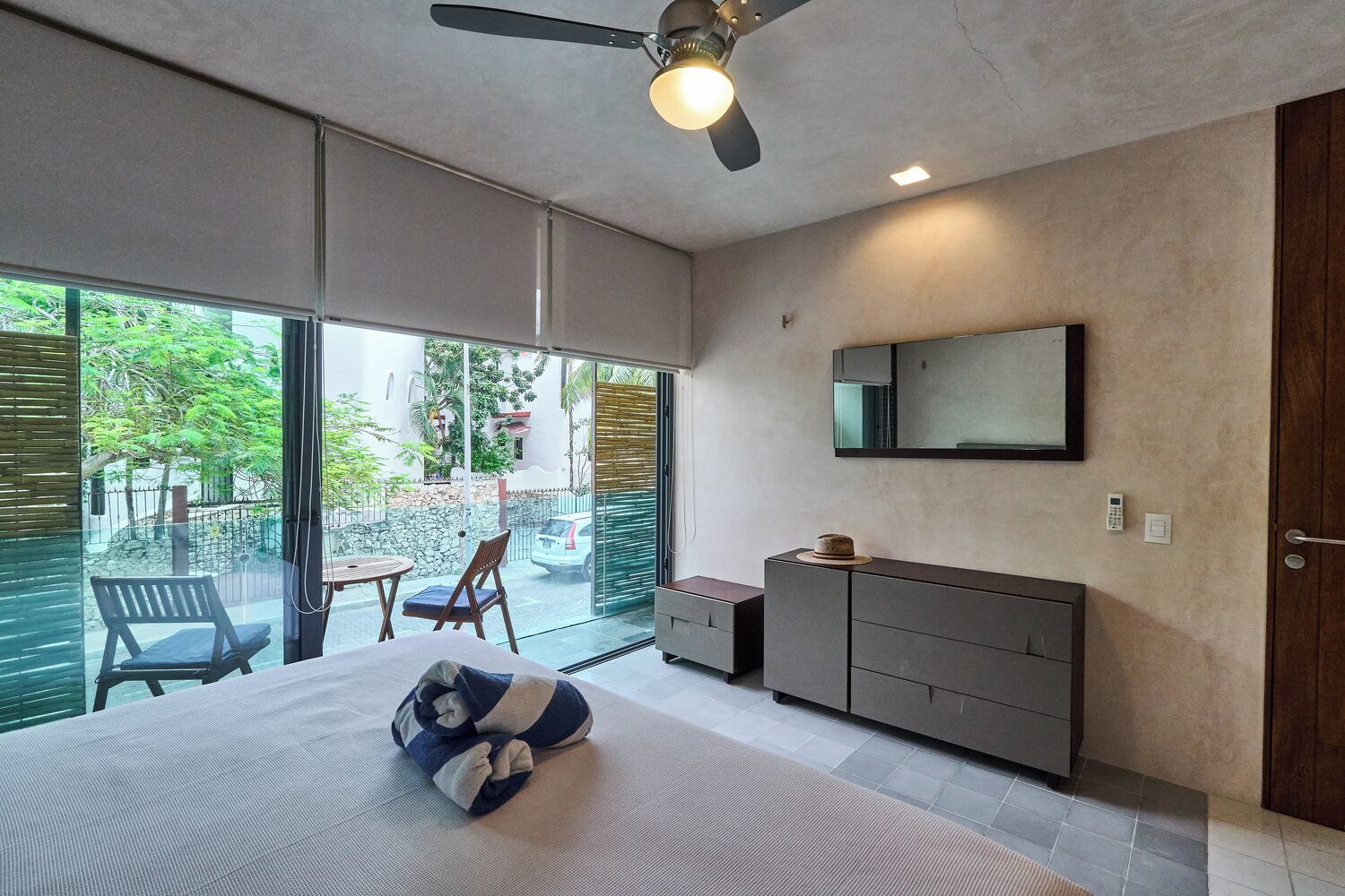Un dormitorio moderno con una cama grande, un ventilador de techo, una puerta corrediza de vidrio que da a un balcón y un tocador minimalista.