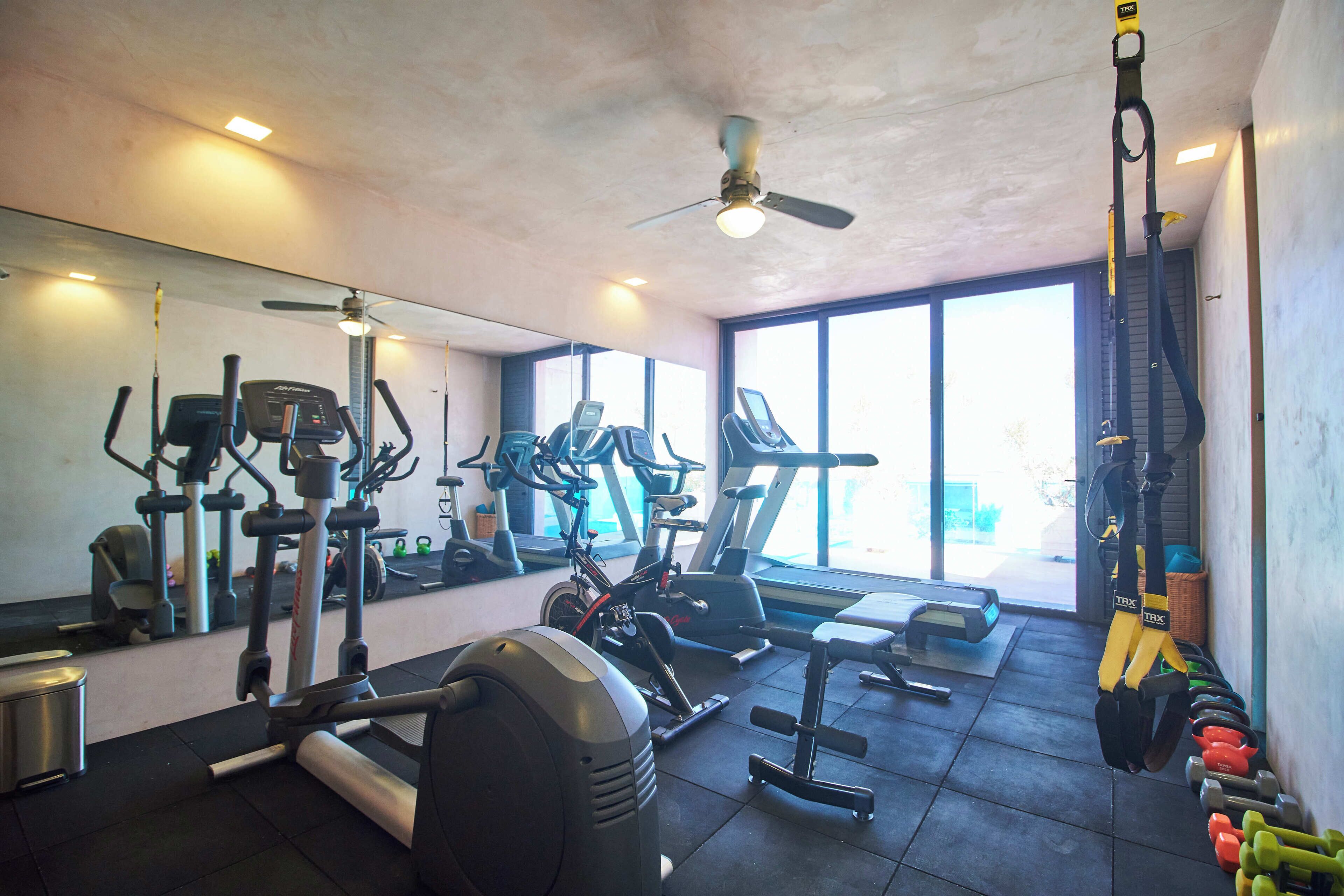 A gym with various exercise equipment, a ceiling fan, and large windows.