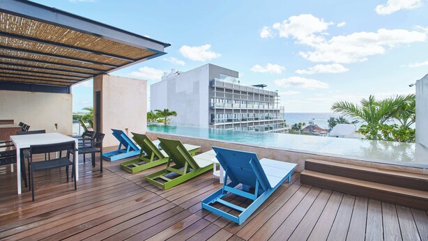 Eine Holzterrasse mit Liegestühlen und einem Tisch, mit Blick auf einen Pool und ein Gebäude, das auf das Meer blickt.