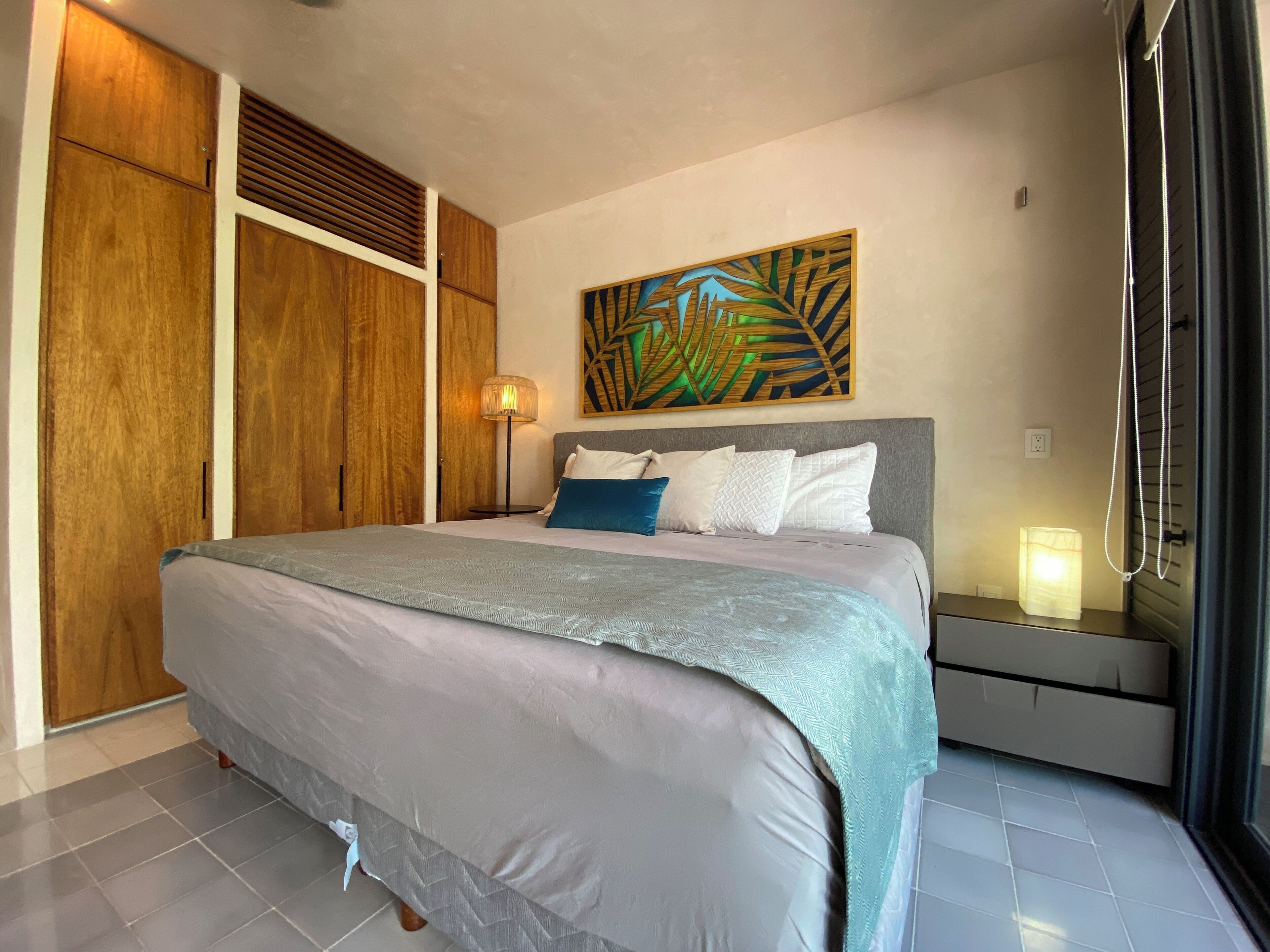 A neatly arranged bedroom with a large bed, a bedside table with a lamp, and a wall-mounted artwork.