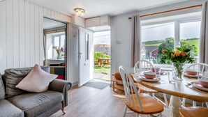 Ett mysigt vardagsrum med en grå soffa, ett matbord för fyra personer och utsikt över en trädgård genom stora fönster.