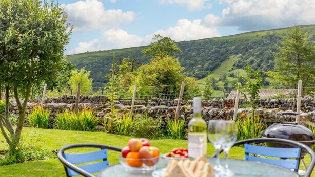 Une table avec un verre de vin, une assiette de fruits et des sandwichs, le tout installé dans un jardin, avec un mur de pierres en arrière-plan et une vue sur des collines ondoyantes.