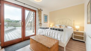 Un dormitorio con un banco de madera, una cama con ropa blanca, una mesita de noche con lámpara y vistas a una terraza de madera y una escalera al exterior.