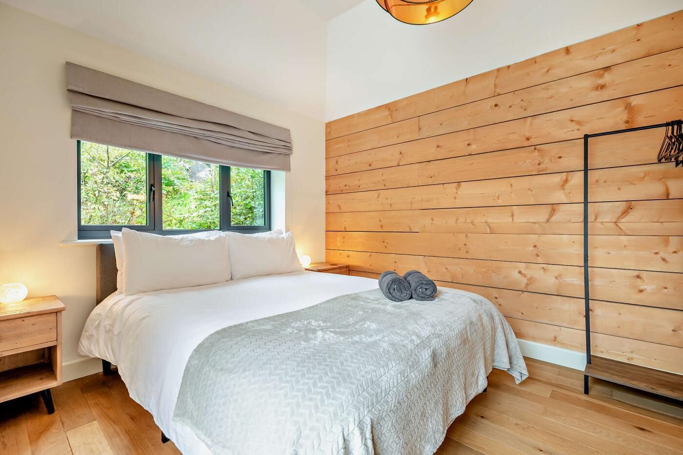 A modern bedroom with a large bed, wooden panel walls, and a window with a view of greenery.