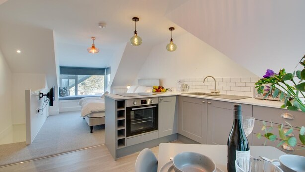 Una cocina moderna con horno empotrado, un comedor con mesa y sillas, y una pequeña zona de estar con una cama.