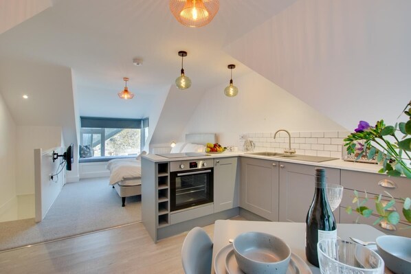 A modern kitchen with a built-in oven, a dining area with a table and chairs, and a small seating area with a bed. - 1 Bed in St. Ives (oc-wrenl) (St Ives)