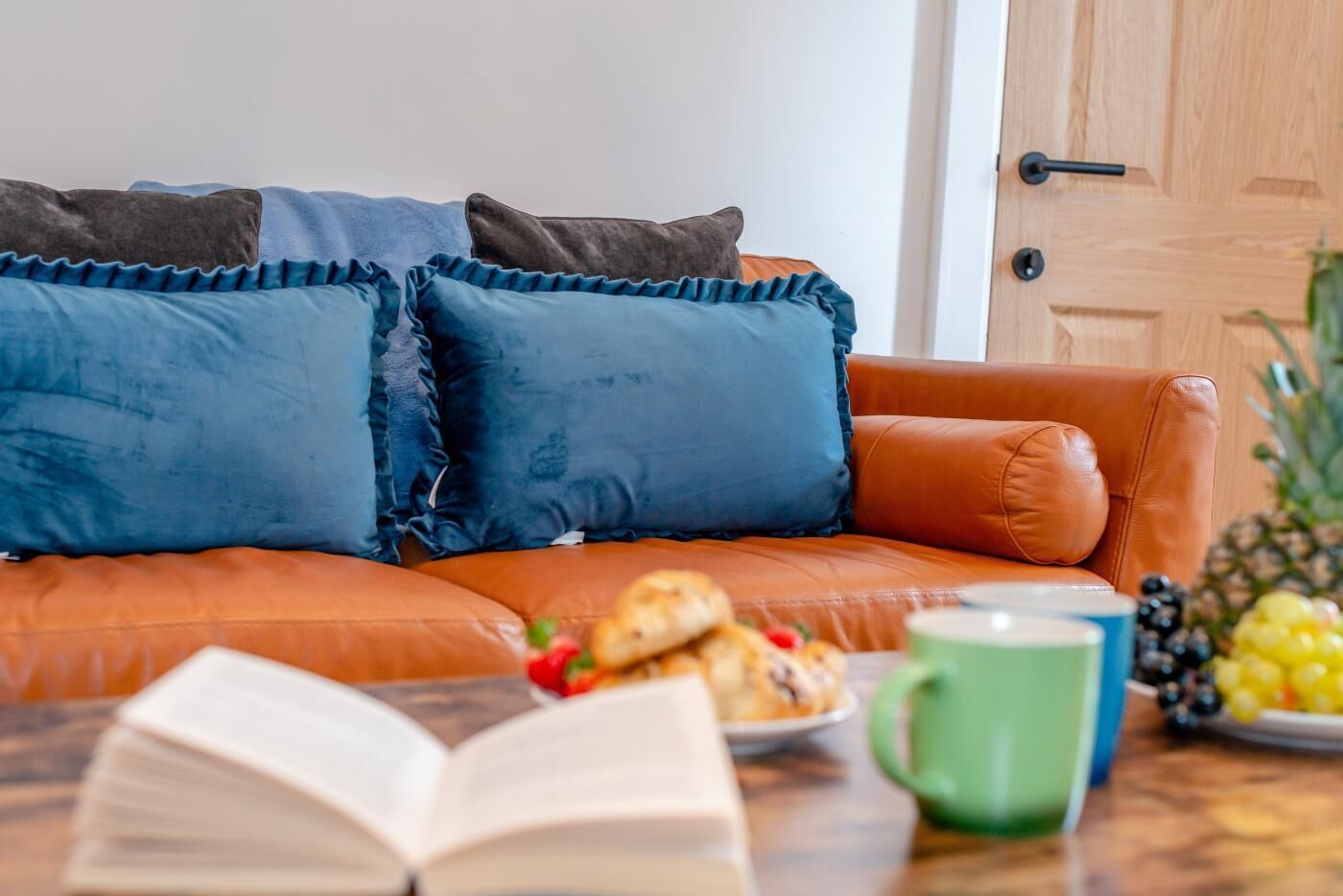 A leather sofa with blue pillows, a book, a plate of pastries, a green mug, and a fruit bowl.