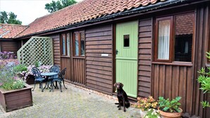 Een houten huis met een groene deur, een terras met een tafel en stoelen, en een hond die bij de deur staat.