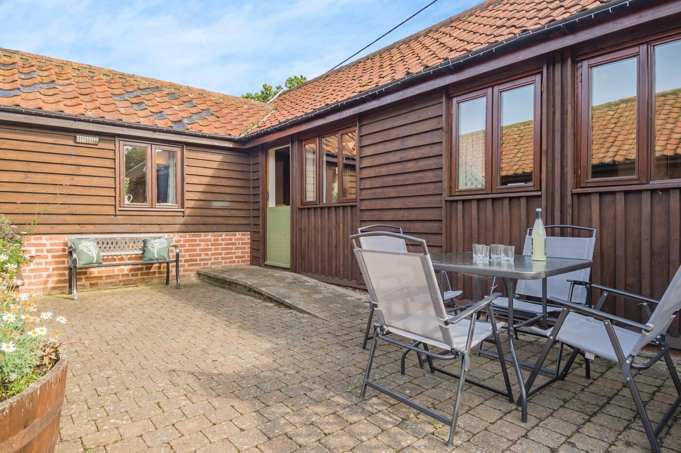 A wooden house with a tiled roof, a patio with a table and chairs, and a bench.