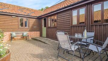 A wooden house with a tiled roof, a patio with a table and chairs, and a bench.