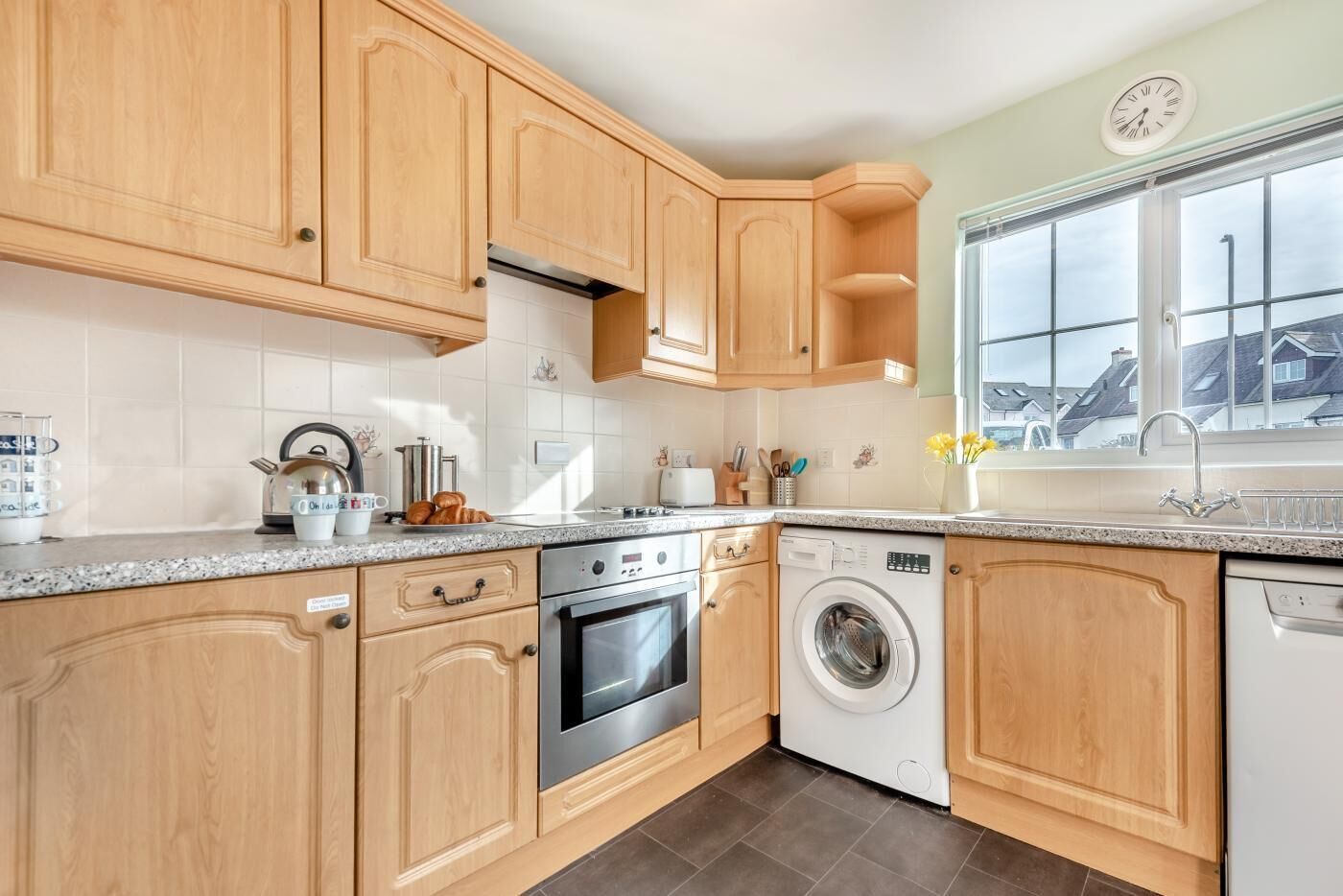Een keuken met houten kastjes, een roestvrijstalen oven, een wasmachine en een raam met uitzicht op huizen.