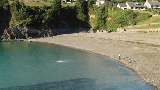 Uma praia costeira com águas claras, um litoral rochoso e casas ao fundo.
