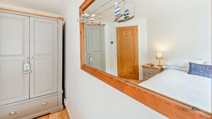 Una camera da letto con testiera in legno, un letto con biancheria da letto bianca, un comodino con una lampada e uno specchio che riflette una porta e una mensola a muro con oggetti decorativi.