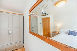 A bedroom with a wooden headboard, a bed with white bedding, a nightstand with a lamp, and a mirror reflecting a door and a wall-mounted shelf with decorative items. - 3 Bed in Little Haven (oc-h27960) (Little Haven)