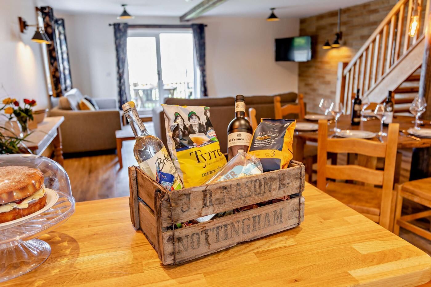 A wooden crate filled with various food items and bottles, placed on a wooden table in a dining area with a staircase and a TV in the background.