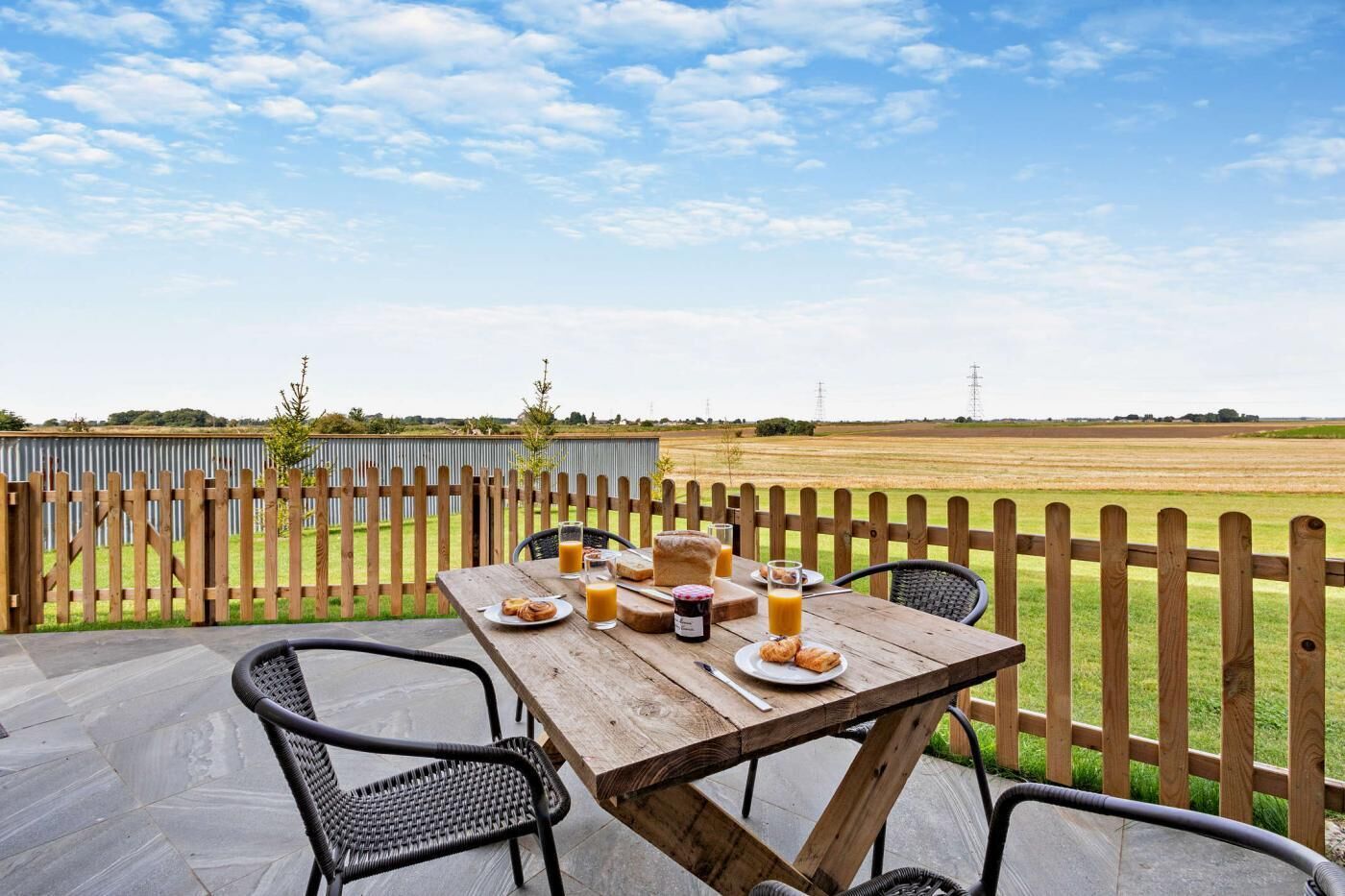 A wooden table with breakfast items and drinks on a patio with a view of a field and power lines.