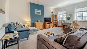 A living room with a blue accent wall, a sofa, a coffee table, a TV, and a dining area with a table and chairs. - 2 Bed in Swanage (oc-s28025) (Swanage)