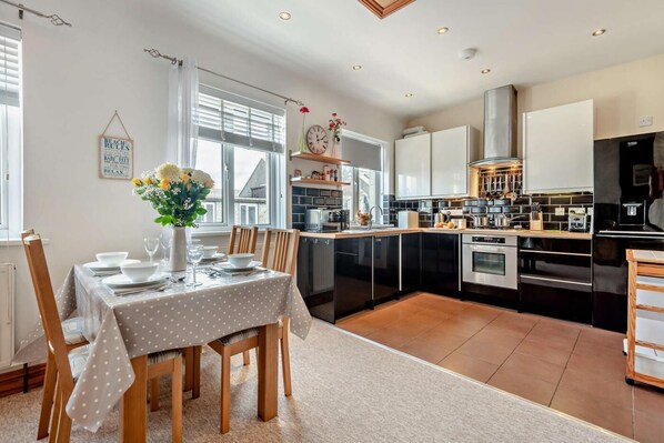 A modern kitchen with black appliances, a dining area with a polka dot tablecloth, and a window with blinds. - 2 Bed in Swanage (oc-s28025) (Swanage)