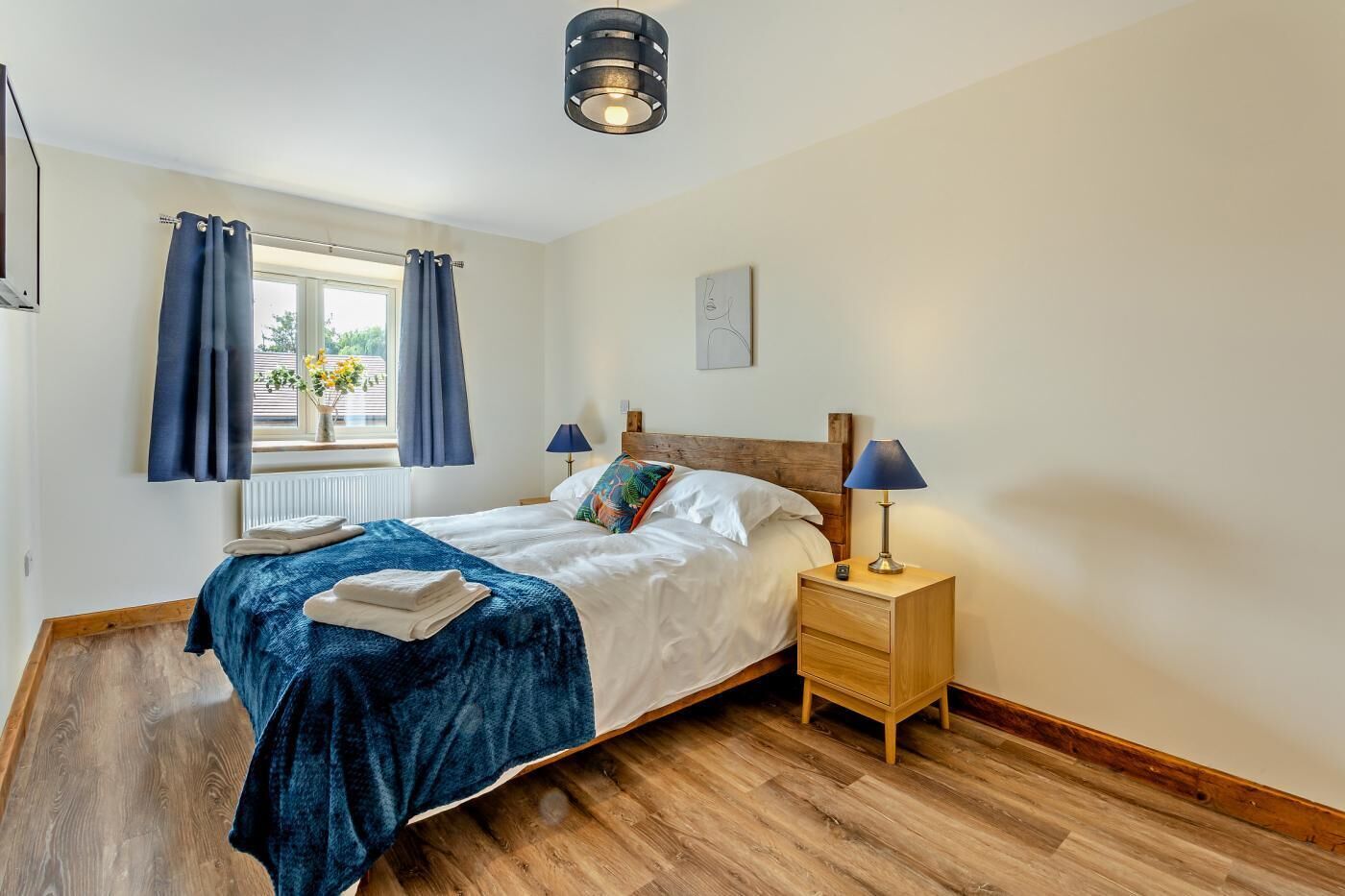 Une chambre avec un plancher en bois, un lit recouvert d’une couverture bleue, une table de chevet avec une lampe et une fenêtre avec des rideaux.