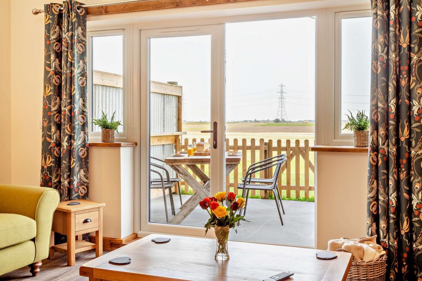 A room with a wooden table, a yellow armchair, and a view of an outdoor patio with a wooden fence and a distant power line.