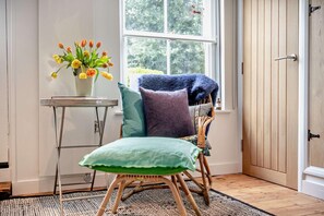 A cozy room with a wicker chair, a small table with a vase of flowers, and a view of greenery through a window. - 1 Bed in Southwold (oc-b27959) (Southwold)
