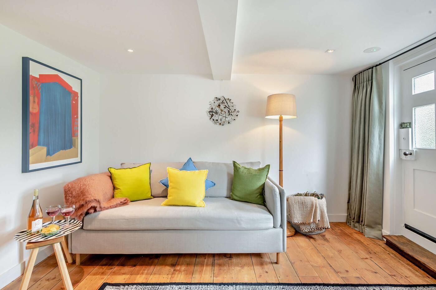 A modern living room with a sofa, colorful cushions, a floor lamp, a painting, and a door with curtains.