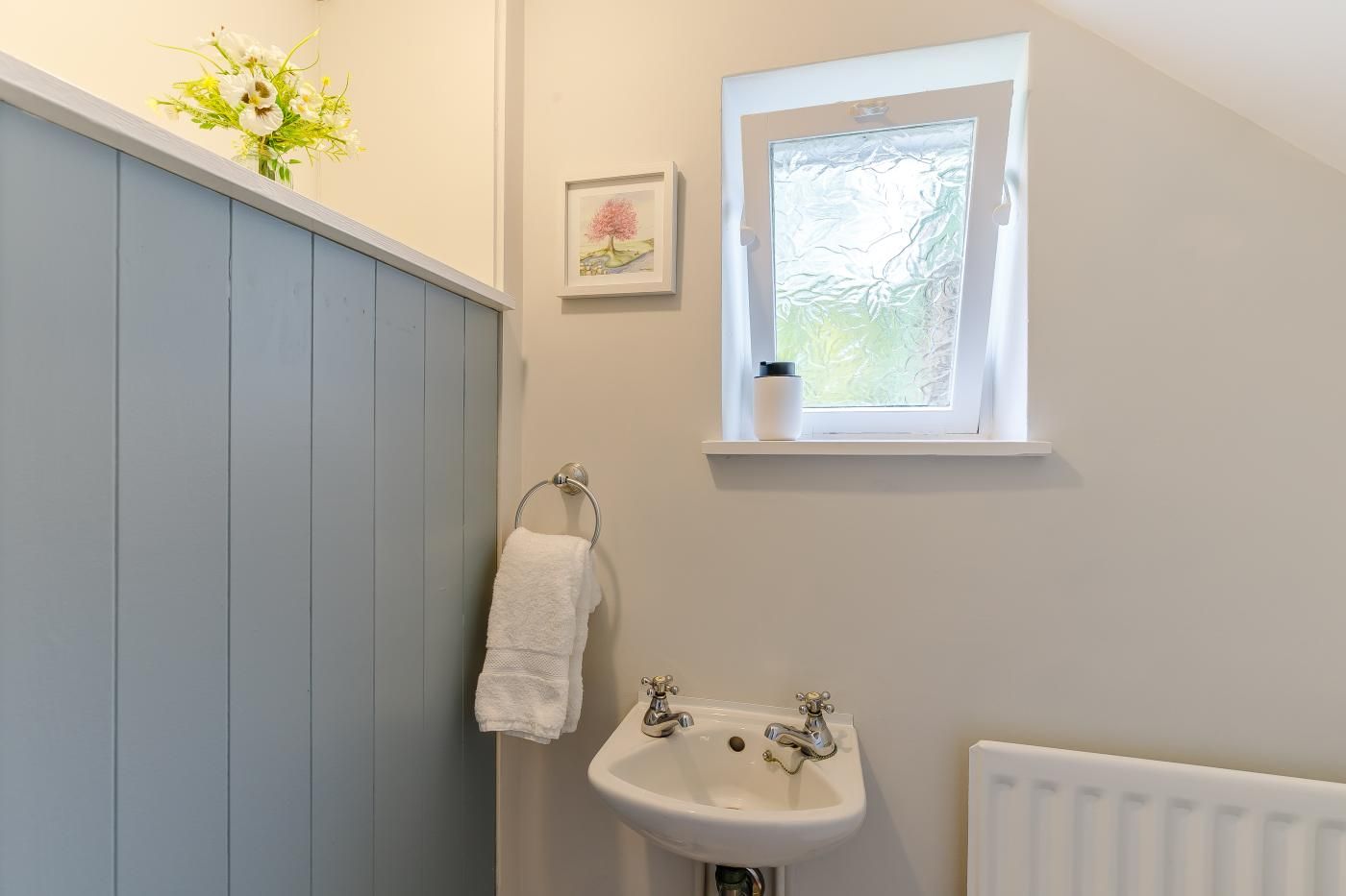 Ein Badezimmer mit weißem Waschbecken, einem Fenster mit Aussicht, einem Handtuchhalter und einem kleinen gerahmten Bild an der Wand.